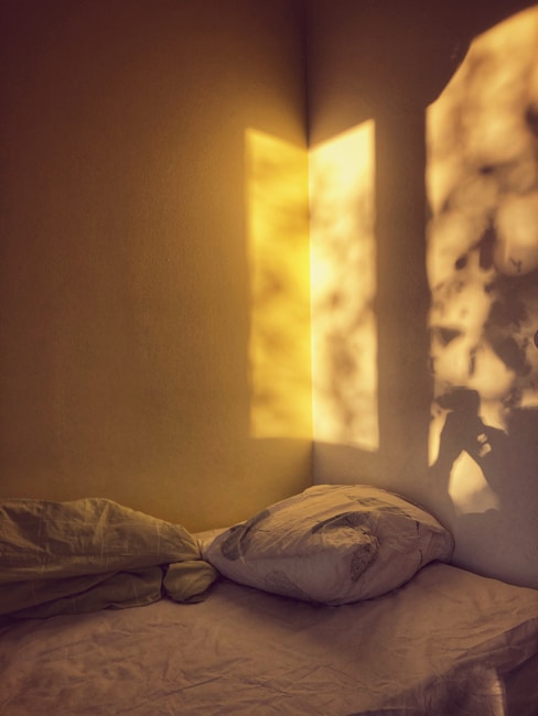 Soft, warm light creates a shadow pattern on the wall, revealing the silhouette of a person holding a camera. A simple bed with a pillow and rumpled blanket is positioned against the corner, suggesting a cozy and serene atmosphere.