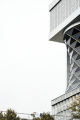 The image features a modern architectural structure with a geometric, lattice-like design. The building has an intricate pattern of repeating shapes on its facade, giving it a contemporary and artistic look. The top corner of the building is visible with a clear, light sky in the background. Trees with sparse foliage are present at the bottom, indicating an urban setting.