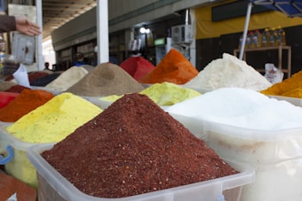 Various containers are filled with colorful powders and spices, arranged in a marketplace setting. The spices are displayed in large, mounded piles within plastic containers. The scene captures a busy market environment with several stalls in the background.