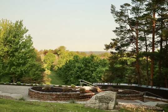 A serene garden landscape featuring lush green trees and a circular, landscaped area with a prominent stone that reads 'Scattering Garden.' The background consists of dense foliage and a clear sky, creating a peaceful and natural scene.