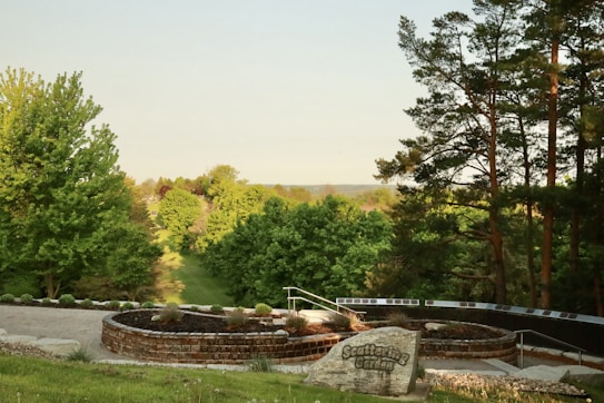 A serene garden landscape featuring lush green trees and a circular, landscaped area with a prominent stone that reads 'Scattering Garden.' The background consists of dense foliage and a clear sky, creating a peaceful and natural scene.