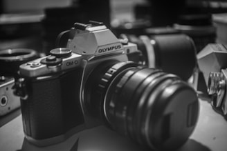 A black and white photograph featuring a vintage-style Olympus camera prominently in the foreground. The camera has a textured grip and multiple lens components. Surrounding the main camera, there are several other camera lenses and equipment slightly out of focus, creating a shallow depth of field effect. The setting appears to be a workspace or a studio environment.