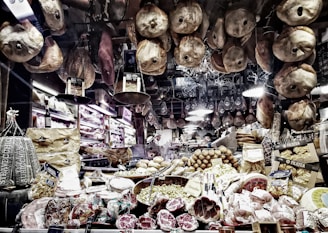 A richly stocked delicatessen displaying an array of cured meats, cheeses, and other gourmet foods. Various types of sausages and hams hang from the ceiling, while wheels of cheese and packaged goods are arranged on the countertop. There are shelves with wine bottles and boxes of pasta, creating a cozy and inviting atmosphere.