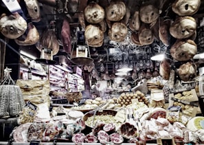 A richly stocked delicatessen displaying an array of cured meats, cheeses, and other gourmet foods. Various types of sausages and hams hang from the ceiling, while wheels of cheese and packaged goods are arranged on the countertop. There are shelves with wine bottles and boxes of pasta, creating a cozy and inviting atmosphere.