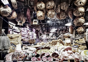 A richly stocked delicatessen displaying an array of cured meats, cheeses, and other gourmet foods. Various types of sausages and hams hang from the ceiling, while wheels of cheese and packaged goods are arranged on the countertop. There are shelves with wine bottles and boxes of pasta, creating a cozy and inviting atmosphere.