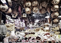 A richly stocked delicatessen displaying an array of cured meats, cheeses, and other gourmet foods. Various types of sausages and hams hang from the ceiling, while wheels of cheese and packaged goods are arranged on the countertop. There are shelves with wine bottles and boxes of pasta, creating a cozy and inviting atmosphere.