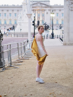 A woman in a white dress and white sneakers is walking by a fenced area. She has a mustard-colored jacket draped over her arm and is looking back towards the camera. The setting appears to be a historic building with large columns and statues in the background.
