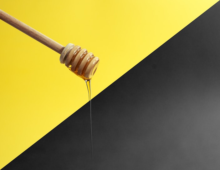 A wooden honey dipper drizzles honey against a background divided diagonally between bright yellow and dark gray.