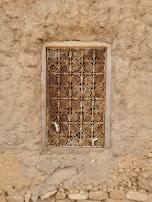 An intricately designed metal grill with spiral patterns covers a small window set into a rough, earthy wall. The frame is rectangular, with weathered wood surrounding the metal work, which displays a mix of rusted and golden hues. The wall is made of uneven, textured material with small stones embedded within it.