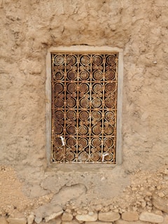 An intricately designed metal grill with spiral patterns covers a small window set into a rough, earthy wall. The frame is rectangular, with weathered wood surrounding the metal work, which displays a mix of rusted and golden hues. The wall is made of uneven, textured material with small stones embedded within it.