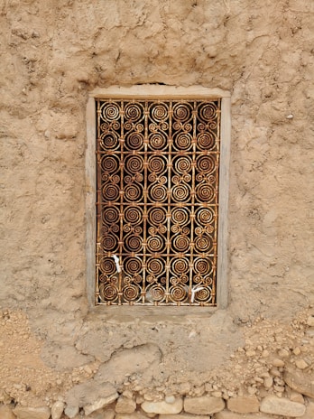 An intricately designed metal grill with spiral patterns covers a small window set into a rough, earthy wall. The frame is rectangular, with weathered wood surrounding the metal work, which displays a mix of rusted and golden hues. The wall is made of uneven, textured material with small stones embedded within it.