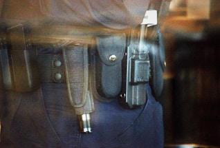 A close-up view of a police or security officer's duty belt equipped with various gear including a baton, a radio, and handcuffs. The equipment is secured in pouches and holsters on a dark-colored fabric belt.