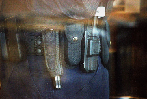 A close-up view of a police or security officer's duty belt equipped with various gear including a baton, a radio, and handcuffs. The equipment is secured in pouches and holsters on a dark-colored fabric belt.