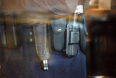 A close-up view of a police or security officer's duty belt equipped with various gear including a baton, a radio, and handcuffs. The equipment is secured in pouches and holsters on a dark-colored fabric belt.