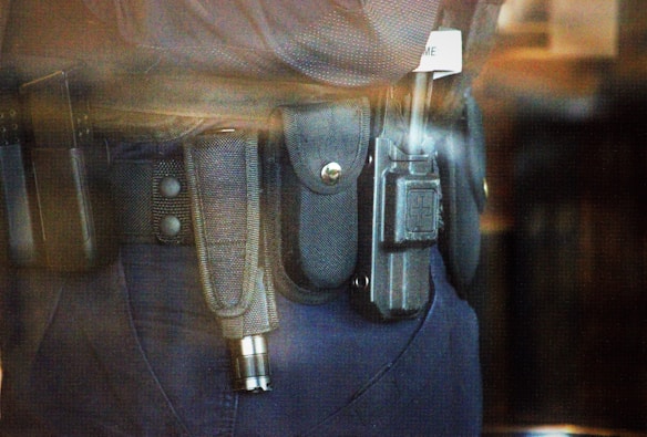 A close-up view of a police or security officer's duty belt equipped with various gear including a baton, a radio, and handcuffs. The equipment is secured in pouches and holsters on a dark-colored fabric belt.