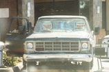 A vintage pickup truck is parked outside a building, with 'FOR SALE' signs visible on its windshield. The truck's driver-side door is open, and there is a shopping cart nearby. The background includes a brick wall and glass doors, suggesting the entrance of a commercial establishment.