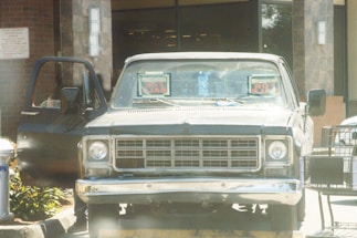 A vintage pickup truck is parked outside a building, with 'FOR SALE' signs visible on its windshield. The truck's driver-side door is open, and there is a shopping cart nearby. The background includes a brick wall and glass doors, suggesting the entrance of a commercial establishment.