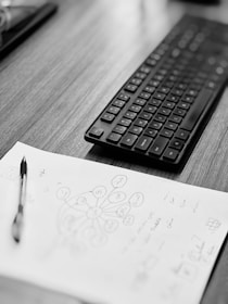 A black and white image showing a desk with a keyboard and a piece of paper containing handwritten notes and a diagram. A pen rests on the paper, and the surface appears to be made of wood.