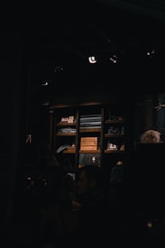 A dimly lit room with wooden shelves stacked with neatly folded clothes and decorative items. People are gathered in the lower part of the image, partially obscured by darkness. The focus is on the shelves with warm lighting highlighting the textures of the clothing and objects.