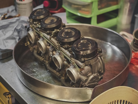 A metal tray on a workbench holding a complex mechanical component with multiple circular elements, likely part of a carburetor system. The background features various tools and containers, suggesting a workshop environment.