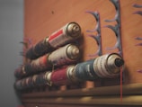 Several spray paint cans are mounted on a wooden wall, held in place by semi-circular holders. The arrangement shows a variety of colors and brands.