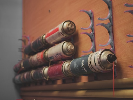 Several spray paint cans are mounted on a wooden wall, held in place by semi-circular holders. The arrangement shows a variety of colors and brands.