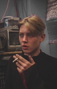 A young person with blonde hair is holding a cigarette close to their lips. The individual is indoors, set against a cluttered background which includes electronic equipment and storage containers. The lighting is dim, creating a moody atmosphere.
