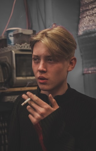 A young person with blonde hair is holding a cigarette close to their lips. The individual is indoors, set against a cluttered background which includes electronic equipment and storage containers. The lighting is dim, creating a moody atmosphere.