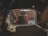 A rectangular rear-view mirror shows a reflection of a person holding a camera. The mirror has dust and smudges on its surface, and the background is a bit blurry with indistinguishable objects.