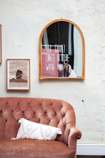 A vintage-style interior features a brown tufted sofa with a textured white pillow. Above the sofa hangs a rectangular art print and an arched mirror with the words 'BAR CHAPEL LOOKS BETTER WITH YOU' written on it. A person is partially visible via the mirror reflection, holding a camera.