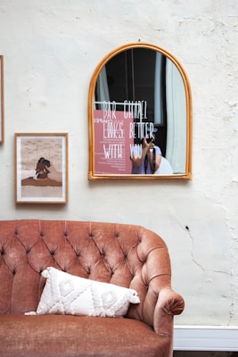 A vintage-style interior features a brown tufted sofa with a textured white pillow. Above the sofa hangs a rectangular art print and an arched mirror with the words 'BAR CHAPEL LOOKS BETTER WITH YOU' written on it. A person is partially visible via the mirror reflection, holding a camera.