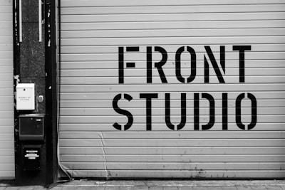 A large garage door features bold, industrial-style lettering spelling 'FRONT STUDIO'. To the left, a wall includes a box labeled 'WORKS OF FICTION'. The color scheme is monochromatic, with a focus on contrasting black and white.