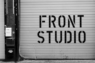 A large garage door features bold, industrial-style lettering spelling 'FRONT STUDIO'. To the left, a wall includes a box labeled 'WORKS OF FICTION'. The color scheme is monochromatic, with a focus on contrasting black and white.