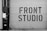 A large garage door features bold, industrial-style lettering spelling 'FRONT STUDIO'. To the left, a wall includes a box labeled 'WORKS OF FICTION'. The color scheme is monochromatic, with a focus on contrasting black and white.