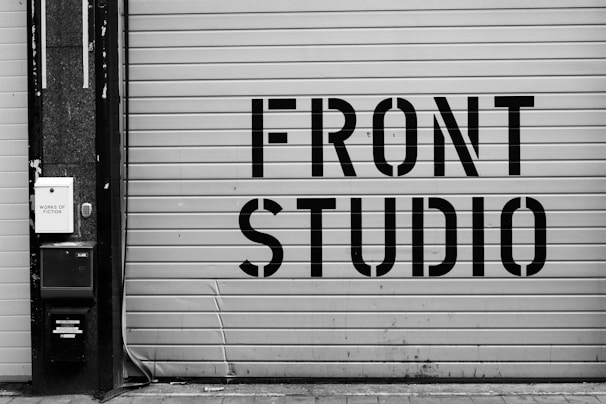 A large garage door features bold, industrial-style lettering spelling 'FRONT STUDIO'. To the left, a wall includes a box labeled 'WORKS OF FICTION'. The color scheme is monochromatic, with a focus on contrasting black and white.
