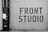 A large garage door features bold, industrial-style lettering spelling 'FRONT STUDIO'. To the left, a wall includes a box labeled 'WORKS OF FICTION'. The color scheme is monochromatic, with a focus on contrasting black and white.