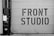 A large garage door features bold, industrial-style lettering spelling 'FRONT STUDIO'. To the left, a wall includes a box labeled 'WORKS OF FICTION'. The color scheme is monochromatic, with a focus on contrasting black and white.