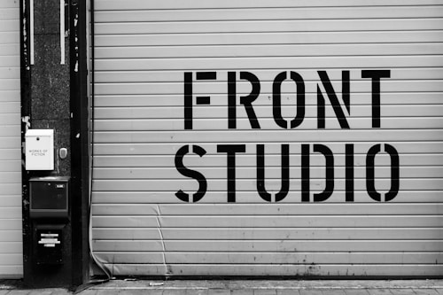 A large garage door features bold, industrial-style lettering spelling 'FRONT STUDIO'. To the left, a wall includes a box labeled 'WORKS OF FICTION'. The color scheme is monochromatic, with a focus on contrasting black and white.
