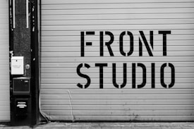 A large garage door features bold, industrial-style lettering spelling 'FRONT STUDIO'. To the left, a wall includes a box labeled 'WORKS OF FICTION'. The color scheme is monochromatic, with a focus on contrasting black and white.