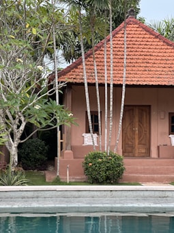 A small, quaint house with a red tiled roof is nestled among lush tropical vegetation. The wooden door is framed by greenery, and two white chairs sit on the porch, hinting at a peaceful and relaxed atmosphere. The foreground features a clear pool, adding a refreshing blue contrast to the earthy tones of the structure.