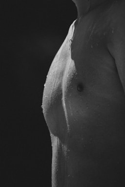 A monochrome close-up of a person's torso, showing water droplets on the skin. The lighting creates contrast and highlights the texture and definition of the body.