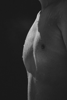 A monochrome close-up of a person's torso, showing water droplets on the skin. The lighting creates contrast and highlights the texture and definition of the body.