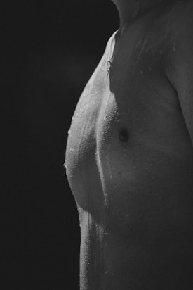 A monochrome close-up of a person's torso, showing water droplets on the skin. The lighting creates contrast and highlights the texture and definition of the body.