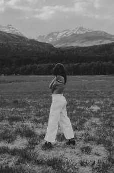 A person standing in a vast field with a mountainous backdrop, wearing white pants and no shirt. The scene is captured in black and white, emphasizing the natural landscape and the person's pose.