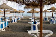 A beach scene with straw umbrellas evenly spaced in rows on a sandy shore. The sun loungers are stacked and placed beneath each umbrella, while gentle waves can be seen in the background along with a hint of the blue sea.