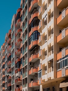 Tall residential buildings with multiple stories, featuring balconies with red railings and glass panels. The facade of the buildings consists of beige and orange segments. Air conditioning units are visible on some balconies.