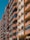 Tall residential buildings with multiple stories, featuring balconies with red railings and glass panels. The facade of the buildings consists of beige and orange segments. Air conditioning units are visible on some balconies.
