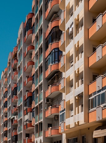 Tall residential buildings with multiple stories, featuring balconies with red railings and glass panels. The facade of the buildings consists of beige and orange segments. Air conditioning units are visible on some balconies.