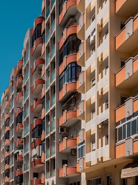 Tall residential buildings with multiple stories, featuring balconies with red railings and glass panels. The facade of the buildings consists of beige and orange segments. Air conditioning units are visible on some balconies.