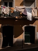 Laundry hangs from a balcony railing, casting shadows on the yellow wall of a building. Various fabrics and towels of different colors and patterns are draped over the railing. Below, two dark green doors provide a contrast to the sunlit textiles above.